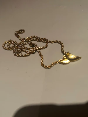 Guldhalsband med hjärta och vinge - Snyggt halsband i guldfärgad metall med tvinnad kedja och ett hängsmycke i form av ett hjärta och en vinge. Perfekt accessoar för dig som gillar detaljer med symbolik. Passar till många olika stilar och är lätt att matcha med andra smycken. Nypris: 350kr