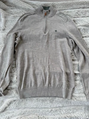 Beige stickad tröja från Morris - Snygg beige stickad tröja från Morris i 100% merinoull. Tröjan har halv dragkedja med läderdetalj, hög krage och diskret logga broderad på bröstet. Perfekt för dig som gillar stilrena och klassiska plagg.