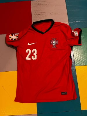 Portugal matchtröja Nike #23 Vitinha - Röd Portugal matchtröja från Nike med nummer 23 och namnet Vitinha på ryggen. Tröjan har svarta detaljer vid kragen, landslagets emblem på bröstet och officiella turneringsmärken på ärmarna. Tillverkad i lätt och ventilerande material, perfekt för fotboll.