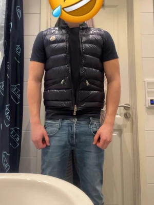 Mörkblå dunväst från Moncler -  Mörkblå dunväst från Moncler med glansig finish och hög krage. Västen har två dragkedjeförsedda fickor framtill och Moncler-logga på bröstet. Perfekt att bära över en t-shirt för en trendig look.