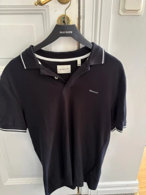 Svart pikétröja från GANT - Klassisk svart pikétröja från GANT med vit kant på krage och ärmslut. Tröjan har korta ärmar, knappslå och diskret GANT-logga broderad på bröstet. Tillverkad i mjuk bomull som känns skön mot huden.