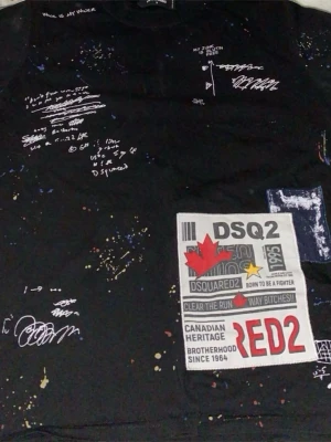 Svart DSquared2 t-shirt med tryck - Cool svart t-shirt från DSquared2 med vita handskriftsliknande tryck och färgstänk över hela tröjan. Stor patch med DSQ2 och röda detaljer framtill. T-shirten har klassisk passform och rund hals. Perfekt för dig som gillar streetwear och statement-plagg.