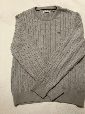 Ljusgrå kabelstickad tröja GANT - Snygg ljusgrå kabelstickad tröja från GANT med rund halsringning och diskret logga broderad på bröstet. Tröjan har ribbade muddar vid ärmslut och nederkant. Perfekt för dig som gillar klassisk och stilren look. Storlek 158/164.