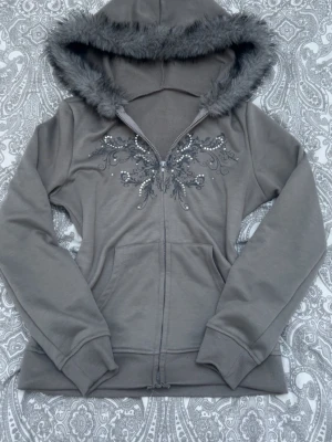 Grå hoodie med fuskpäls och pärlor - Snygg grå hoodie med dragkedja, dekorerad med pärlor och tryck framtill. Luva med päls, supersnygg och mysig, nästan aldrig använd (max 2-3 gånger), säljer eftersom att jag aldrig använder