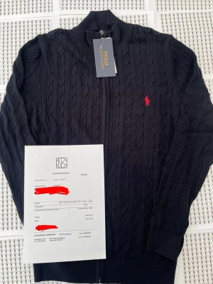 Svart kabelstickad ziptröja Polo Ralph Lauren - Kabelstickad svart ziptröja från Polo Ralph Lauren med hög krage och ribbade muddar. Tröjan har det klassiska röda broderade logot på bröstet och är tillverkad i mjukt material. Perfekt för dig som gillar stilrena och tidlösa plagg.