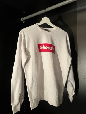 Vit sweatshirt med röd Sheesh-tryck - Vit sweatshirt med ett stort rött Sheesh-tryck på bröstet. Klassisk rund halsringning och långa ärmar. Tillverkad i mjuk bomull för en bekväm och avslappnad stil. Perfekt för dig som vill ha en enkel men trendig look.