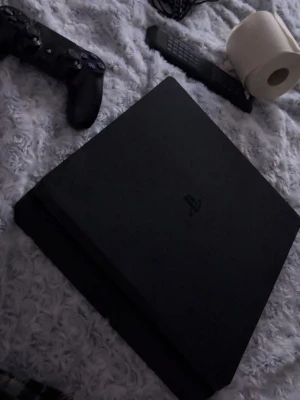 PlayStation 4 Slim spelkonsol svart - Säljer en svart PlayStation 4 Slim från Sony. Konsolen har en stilren, matt finish och den klassiska fyrkantiga formen med rundade kanter. Perfekt för gaming och streaming, passar till alla PS4-spel och har HDMI-utgång. Ingår en kontroll, och två spel nba2k23 samt spiro.