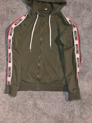 Olivgrön hoodie från Moschino - Snygg olivgrön hoodie från Moschino med dragkedja framtill och vita dragsnören i huvan. Längs ärmarna finns vita band med Moschino-logga i svart. Tillverkad i mjukt bomullsmaterial, perfekt för en avslappnad och trendig look.