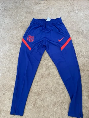 Blå FC Barcelona träningsbyxor Nike - Snygga blå träningsbyxor från Nike med FC Barcelona-logga och röda detaljer. Byxorna har smal passform, resår i midjan och dragkedjor vid bensluten. Perfekta för fotbollsträning eller chill. Materialet är troligtvis polyester.