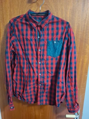 Rödblå rutig skjorta med ficka - Snygg röd och blå rutig skjorta med lång ärm och klassisk krage. Skjortan har en kontrasterande bröstficka i blått tyg och knäppning framtill. Perfekt för en avslappnad och trendig stil.