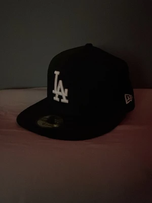 Svart LA New Era keps - Snygg svart keps från New Era med LA-logga broderad i vitt framtill. Klassisk flat brim-modell och broderade detaljer på sidan och bak. Perfekt för dig som gillar streetwear och sportig stil. Storlek 6 & 7/8. Hör av er vid bud och eventuella frågor😊