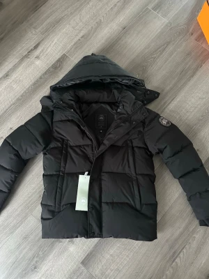 Svart Canada goose jacka  - Snygg svart pufferjacka från Canada goose med stor huva och patch på ärmen. Jackan har breda kanaler, dragkedja och tryckknappar framtill samt två sidofickor. Perfekt för kalla vinterdagar och riktigt trendig look.