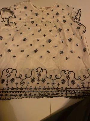 Vit blus med svart brodyr - Söt vit blus med svart broderat mönster och vågig kant längst ner. Blusen har korta ärmar med dekorativ kant och ett luftigt, lätt tyg som ger en avslappnad känsla. Perfekt för dig som gillar boho-vibbar och detaljer.