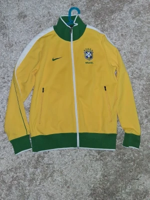 2002 vintage Brasil Nike track jacket (ÄKTA) - Snygg gul track jacket från Nike med Brasil-logga och gröna detaljer. Jackan har hel dragkedja, två fickor med dragkedja och vit rand längs ärmarna. Perfekt för dig som gillar fotboll och vill representera Brasilien. Helt oanvänd och är retro/vintage kofta i tidigt 2000-tale.