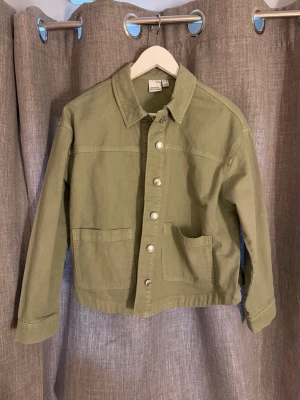 Olivgrön jeansjacka med fickor - Snygg olivgrön jeansjacka med raka linjer och klassisk krage. Köpt för 600kr och är endast använd 1 gång. Jackan har två stora framfickor och en mindre bröstficka, samt silverfärgade knappar framtill. Perfekt för dig som gillar en enkel men trendig look.