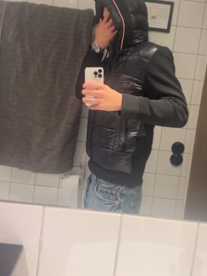 Svart dunjacka med huva från Moncler - Säljer en svart dunjacka från Moncler med huva och quiltad design. Jackan har stickade ärmar och muddar, samt dragkedja framtill. Perfekt för kalla vinterdagar och har en modern, sportig look.