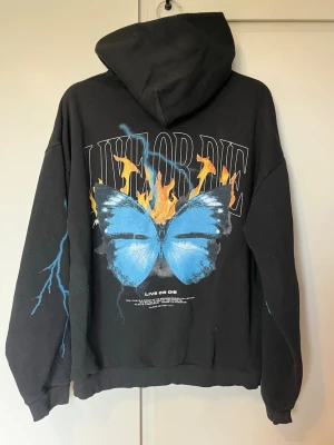 Hoodie - Säljer en svart hoodie med stor blå fjäril och eld på ryggen samt texten 'LIVE OR DIE'. På framsidan står det 'ALONE against ALL' och ärmarna har blå blixtdetaljer. 