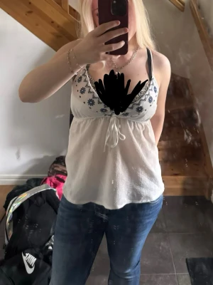 Vit broderad linnetopp  - Söt vit linnetopp  med tunna axelband och blå broderade blommor längs v-ringningen. Toppen har knyt under bysten och är tillverkad i ett lätt, luftigt material som passar perfekt till sommaren. Enkel att matcha med jeans eller kjol. Linnet är aldrig använt men legat i garderoben ett tag. Linnet var tyvärr för liten i bysten
