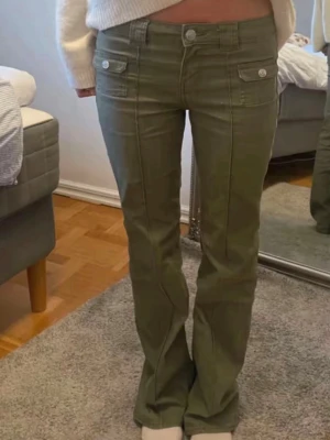 Hm Olivgröna bootcut-byxor - De trendiga olivgröna bootcut-byxorna från Hm . dekorativa fickor framtill och silverfärgade knappar. Byxorna har en klassisk passform med raka ben och markerade sömmar. Perfekta för dig som vill ha en cool och avslappnad look.