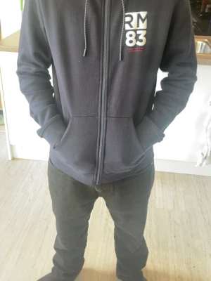 Mörkblå hoodie med dragkedja RM83 - Snygg mörkblå hoodie från RM83 med dragkedja framtill och vit logga på bröstet. Hoodien har huva med dragsko och två stora fickor framtill. Perfekt för en avslappnad och cool stil. Materialet känns mjukt och bekvämt.
