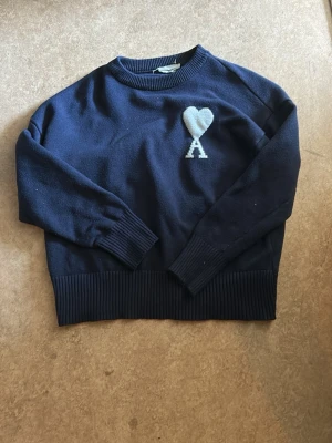 Mörkblå stickad tröja Ami Paris - Mörkblå stickad tröja från Ami Paris med ikoniskt vitt hjärta och bokstaven A broderat på bröstet. Tröjan har rund hals, ribbade muddar och nederkant. Perfekt för dig som gillar stilrena detaljer och klassisk design.