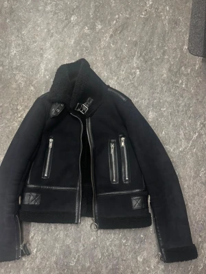 Svart pälsjacka med dragkedjor - Snygg svart pälsjacka med detaljer i läderimitation, hög krage med spänne och flera dragkedjeförsedda fickor framtill. Jackan har en kortare modell och mjukt foder som håller värmen. Perfekt för kalla dagar och ger en cool look.
