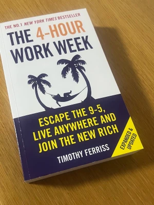 The 4-Hour Work Week pocketbok - En pocketbok med titeln 'The 4-Hour Work Week' av Timothy Ferriss. Omslaget är vitt och mörkblått med orange och gul text samt en illustration av en person i hängmatta mellan två palmer. Boken är på engelska och har mjuka pärmar.