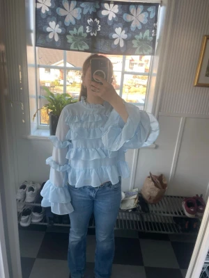 Ljusblå volangblus med lång ärm - Supersöt ljusblå blus med flera lager volanger och transparenta långärmade ärmar. Blusen har en lös passform och rund halsringning. Perfekt för dig som gillar romantisk och lekfull stil. Materialet känns lätt och luftigt.