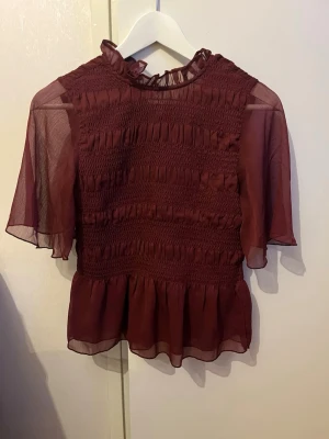 Vinröd blus med volang och smock - Snygg vinröd blus med transparenta, vida ärmar och smockdetaljer över bröstet. Blusen har volang vid halsen och nederkanten samt rundad halsringning. Tillverkad i ett lätt, skirt material som ger en romantisk känsla.