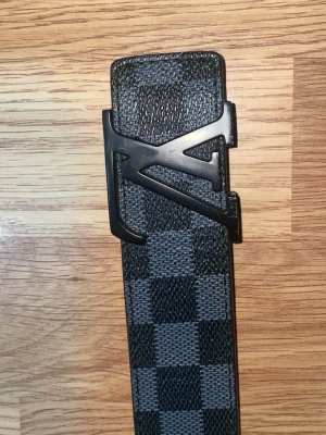 Svart rutigt bälte - Säljer ett svart bälte  med klassiskt rutigt mönster och en stor svart logga i metall som spänne. Bältet har en modern och stilren look, perfekt för att lyfta vilken outfit som helst.