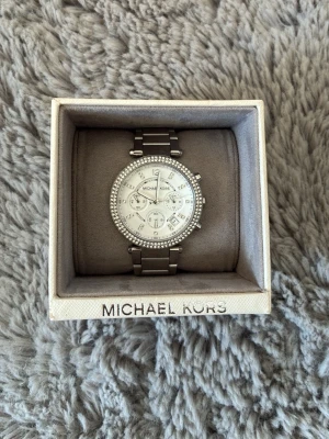 Silverfärgad klocka från Michael Kors - Stilren klocka från Michael Kors i silverfärg med länkarmband och gnistrande stenar runt urtavlan. Urtavlan är vit med flera små visare och tydliga siffror. Perfekt accessoar för dig som vill ha något elegant och trendigt.