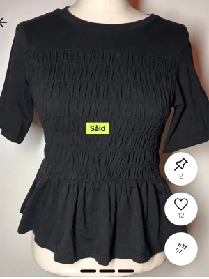 Svart topp med smock och volang - Svart topp med smockad midja och volang nedtill. Modellen har rund halsringning och halvlånga ärmar. Toppens smock ger en snygg och figurnära passform, medan volangen skapar en feminin siluett. Perfekt att styla med jeans eller kjol. Tröjan är samma som jag har, den är tidigare köpt på Plick den är i nytt skick. 