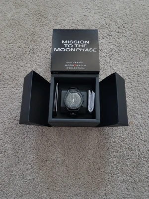 Omega x Swatch MoonSwatch svart klocka - Snygg svart Omega x Swatch MoonSwatch Mission to the Moonphase Snoopy med biokeramiskt material. Klockan har en sportig design med tydlig urtavla, kronograf och svart armband. Perfekt för dig som gillar stilrena accessoarer och ikonisk design.