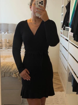 Svart omlottklänning med volang - Svart omlottklänning med v-ringning och långa ärmar. Klänningen har ett knytband i midjan och volangdetalj nedtill. Ribbad struktur ger en snygg och modern look. Perfekt för dig som gillar stilrena och klassiska plagg.