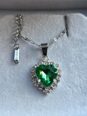 Halsband med grönt hjärthänge - Snyggt halsband i rostfritt stål med ett hjärtformat grönt hänge omgivet av gnistrande klara stenar. Kedjan är silverfärgad i rostfritt stål och har en justerbar längd. Perfekt för dig som vill sticka ut med en färgstark accessoar.