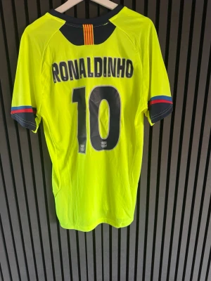 Gul FC Barcelona Ronaldinho tröja - Säljer en gul FC Barcelona matchtröja från Nike med Ronaldinho och nummer 10 på ryggen. Tröjan har klubbmärke på bröstet, svarta detaljer och färgade ränder på ärmsluten. Perfekt för dig som älskar fotboll och klassiska spelare.