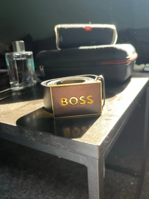 Svart BOSS bälte med gulddetaljer - Snyggt svart bälte från BOSS med en rektangulär metallspänne där BOSS-loggan står i guld. Köpt för 990kr, mest legat still. Bältet har en clean och stilren look som passar till många outfits. Tillverkat i slätt skinn för en exklusiv känsla. Pris kan diskuteras!