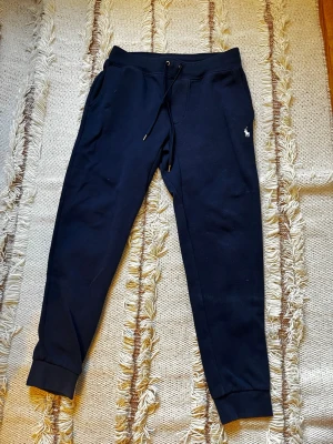 Mörkblå mjukisbyxor från Polo Ralph Lauren - Snygga mörkblå mjukisbyxor från Polo Ralph Lauren med dragsko i midjan och ribbade muddar vid bensluten. Byxorna har fickor på sidorna och en bakficka, samt den klassiska Polo-loggan broderad på vänster sida. Perfekta för chill dagar.
