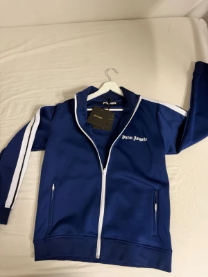 Blå ziptröja från Palm Angels M - Snygg blå ziptröja från Palm Angels med vita detaljer och dragkedja framtill. Tröjan har vita ränder längs ärmarna, ståkrage och två fickor med dragkedja. Perfekt för en sportig och trendig look. Storlek M. Aldrig använd. Helt ny aldrig ens tatt av tagsen. Passar ej mig därför säljer jag istället för att den ska sitta i garderoben. 