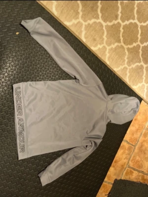 Grå hoodie från Under Armour - Snygg grå hoodie från Under Armour med stor logga längs sidan. Tillverkad i ett mjukt och stretchigt material som känns sportigt och bekvämt. Hoodien har huva och långa ärmar, perfekt för en avslappnad och aktiv stil.