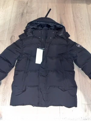 Svart dunjacka från Canada Goose - Snygg svart dunjacka från Canada Goose med huva och klassisk logotyp på ärmen. Jackan har dragkedja och knappar framtill samt flera fickor. Perfekt för kalla vinterdagar och riktigt varm tack vare dunfyllningen.