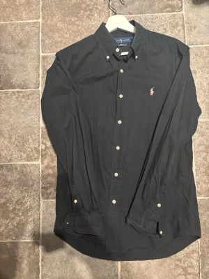 Svart skjorta från Ralph Lauren - Klassisk svart skjorta från Ralph Lauren med rosa broderad logga på bröstet. Skjortan har knappar hela vägen, button-down krage och är tillverkad i bomull. Perfekt för en stilren och enkel look.