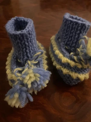 Blå stickade babysockor med tofsar - Supersöta handstickade babysockor i blått och ljusgult. Sockorna har ribbad kant, dekorativa tofsar och snörning runt ankeln. Perfekta för att hålla små fötter varma och mysiga.