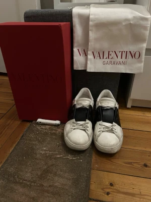 Valentino open Garavani  - Snygga vita sneakers från Valentino open Garavani med svart detalj på sidan. Klassisk låg modell med vita skosnören och gummisula. Kommer med originalkartong och dustbag. Perfekta för dig som vill ha en stilren och exklusiv look. Box, dustbag och skosnören medföljer! Priset är diskuterbart!!