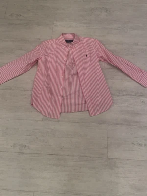 Rosa och vit randig skjorta Ralph Lauren - Klassisk skjorta från Ralph Lauren i rosa och vitt randigt mönster. Skjortan har långa ärmar, knappar framtill och en liten broderad logga på bröstet. Tillverkad i mjuk bomull och har en tidlös, normal passform.