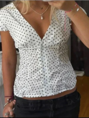 Vit prickig blus med v-ringning - Säljer en vit blus med svart polka dot och djup v-ringning. Blusen har korta ärmar, knappar framtill och figurnära passform. Perfekt för dig som gillar en söt och trendig look. Materialet känns lätt och luftigt.
