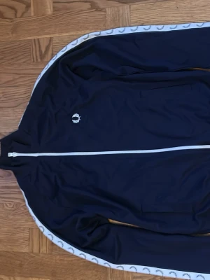 Marinblå Fred Perry track jacket - Snygg marinblå track jacket från Fred Perry med vit dragkedja och vita detaljer längs ärmarna med klassiska Fred Perry-loggor. Jackan har hög krage, ribbade muddar och två sidofickor.