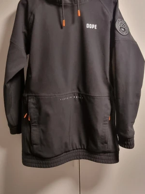Svart anorakjacka från Dope - Svart anorakjacka från Dope med huva, stor magficka och dragkedjor med orange detaljer. Jackan har tryckt logga på bröstet och patch på ärmen. Tillverkad i slitstarkt material, perfekt för kalla dagar.