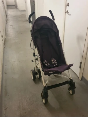 Lila sulky  - En smidig sulky i lila tyg med svart handtag och vita hjul. Vagnen har enkel design, säkerhetsbälte och praktisk fotrem. Perfekt för snabba ärenden eller resor när du behöver en kompakt barnvagn.