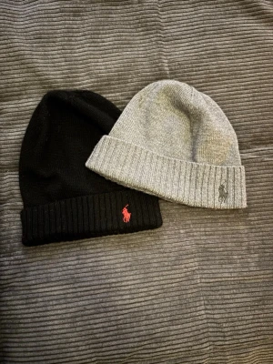 Två mössor från Polo Ralph Lauren - Säljer två stilrena mössor från Polo Ralph Lauren. En svart med röd logga och en grå med mörk logga, båda i mjukt stickat material. Klassisk ribbad kant och broderad logga framtill. Perfekta för kalla dagar och snygga till alla outfits.